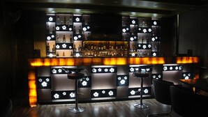 Un área de bar con piso de madera oscura, taburetes y estantes con botellas y vasos.