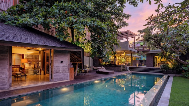 A swimming pool with a patio area, a covered seating area, and a building with an open entrance. - Jadine Bali Villa by Nagisa Bali (Canggu)