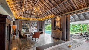 A spacious room with a large bed, a television, a seating area with a chair and a table, and a view of a pool and greenery outside. - Jadine Bali Villa by Nagisa Bali (Canggu)