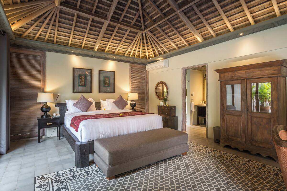 A bedroom with a bed, bedside tables, a bench, a wardrobe, and a ceiling with wooden beams.