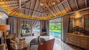 A spacious indoor area with a high ceiling, a pool visible through glass doors, a sofa, a TV, and a wooden cabinet. - Jadine Bali Villa by Nagisa Bali (Canggu)