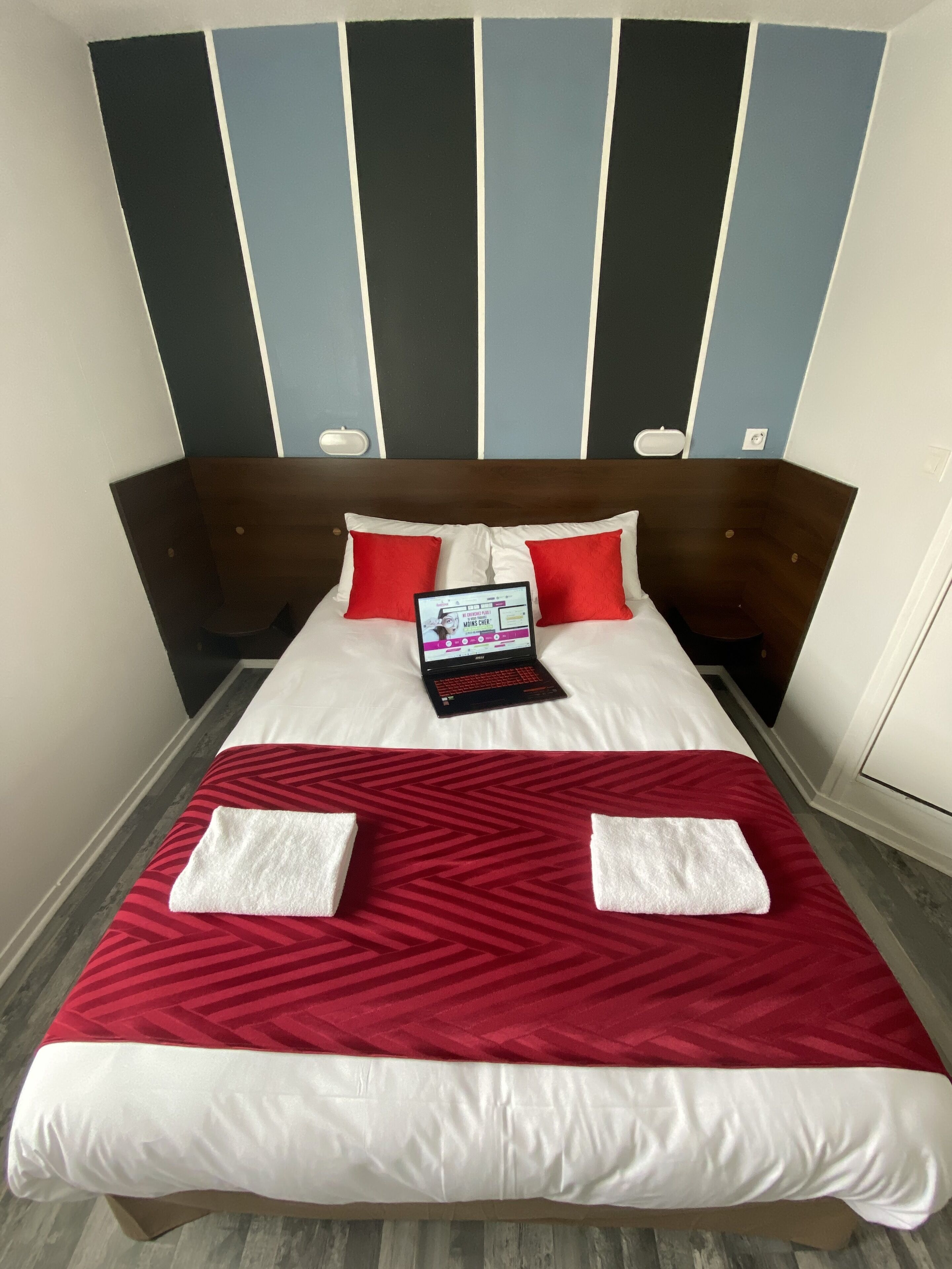 Un lit bien fait, avec un ordinateur portable posé dessus, une literie rouge et blanche, et un mur à rayures rouges et blanches en arrière-plan.
