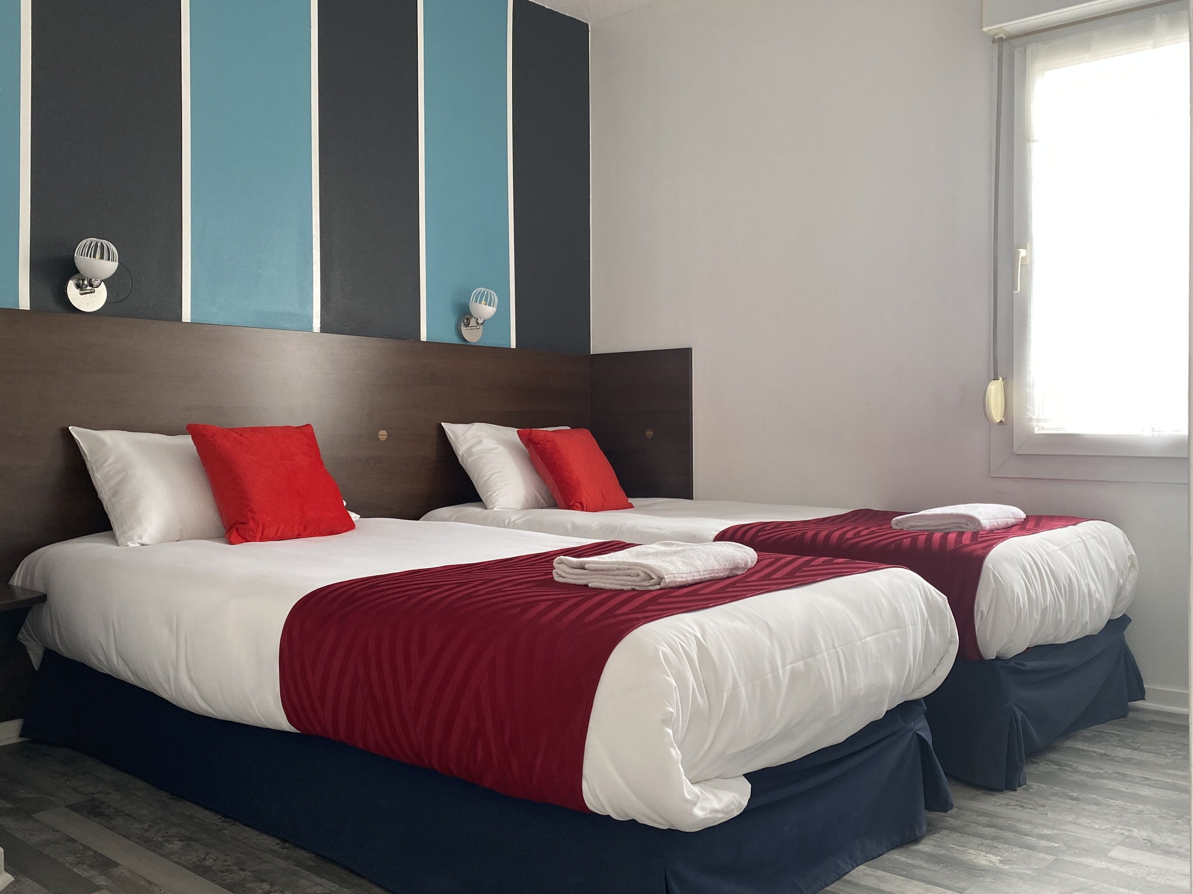 Habitación de hotel con dos camas, cada una con ropa de cama blanca y roja, cabecero oscuro y ventana con cortinas.