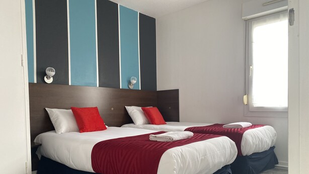 Quarto de hotel com duas camas, cada uma com roupa de cama branca e vermelha, cabeceira escura e design de parede listrado.