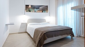 Un dormitorio moderno con una cama grande, una mesita de noche, un cuadro en la pared y una ventana con cortinas translúcidas.