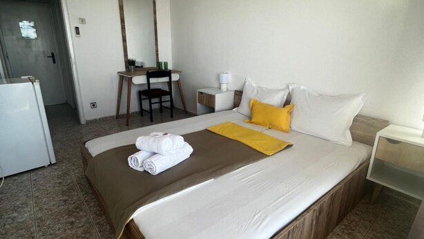 A hotel room with a large bed, a desk with a chair, a small table with a lamp, and an air conditioning unit on the wall. - Sunny Varshava Hotel (Golden Sands)