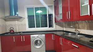 A compact kitchen with red cabinets, a stainless steel washing machine, a gas stove, and a window with blinds. - Apartment 3 Rooms city center Marmoucha (Fes)