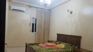 A bedroom with a bed, a nightstand, an air conditioning unit, a window with curtains, and a chandelier. - Apartment 3 Rooms city center Marmoucha (Fes)