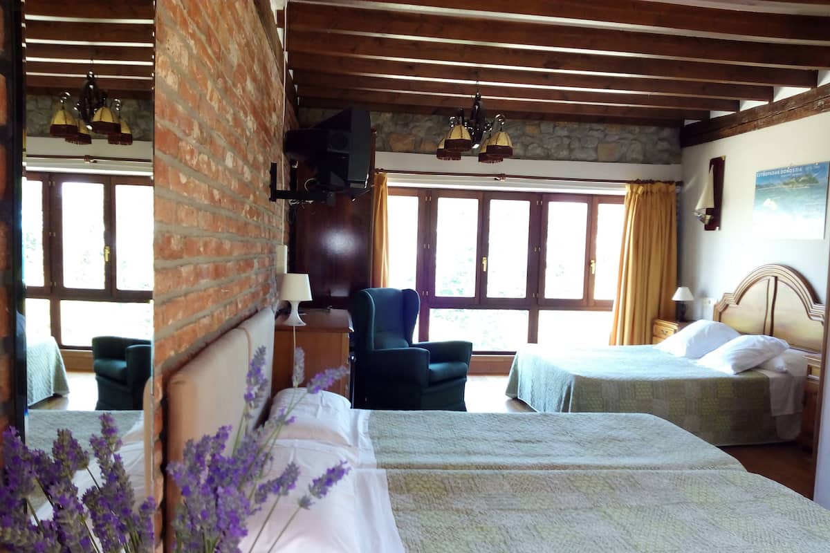 Quarto de hotel com duas camas, parede de tijolos, vigas de madeira, parede de pedra e janelas amplas.