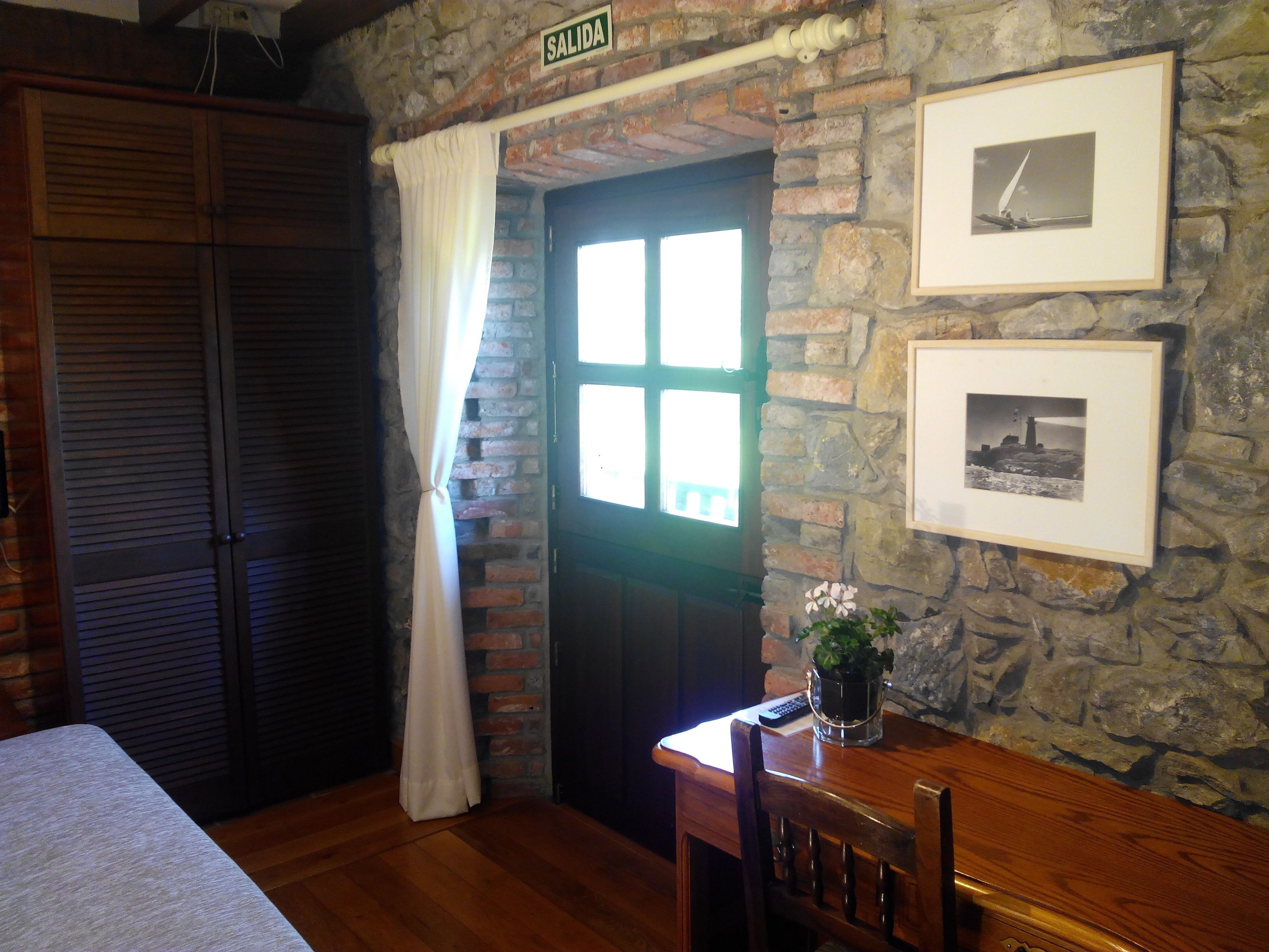 Una habitación con pared de piedra, puerta de madera con ventanal, cama, una mesa con una planta y dos cuadros enmarcados.