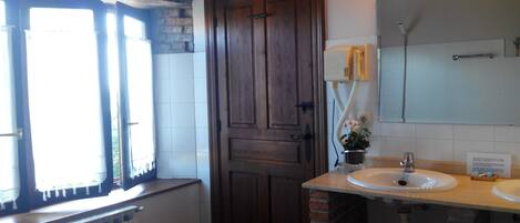Un baño con puerta de madera, ventana con cortinas, radiador, lavabo, espejo y una planta en maceta.