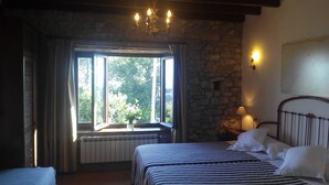 A room with a bed, a window with curtains, a stone wall, a chandelier, and a lamp.
