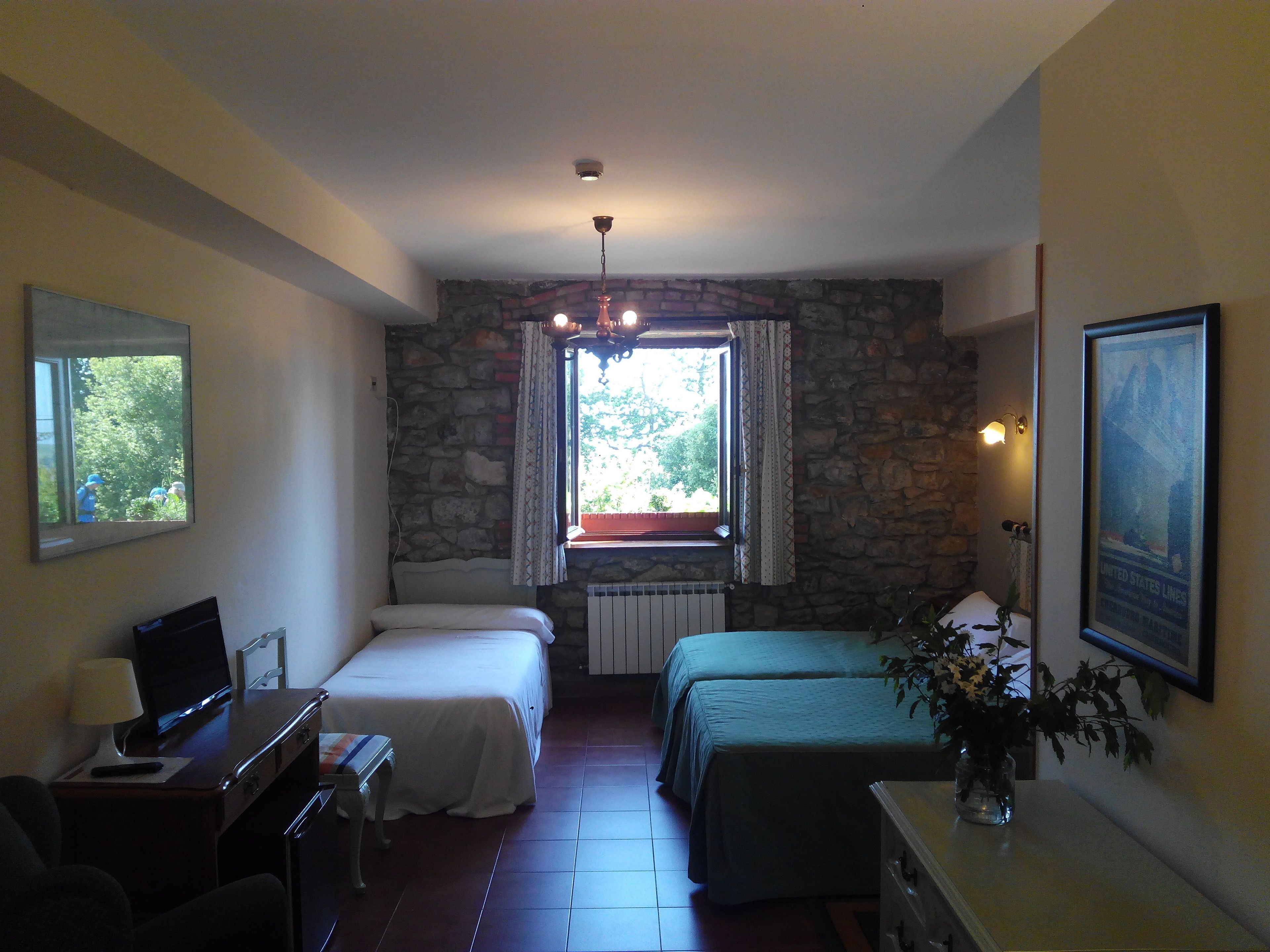 Una habitación con dos camas, un escritorio con computadora, una ventana con cortinas, un muro de piedra y un cuadro enmarcado en la pared.