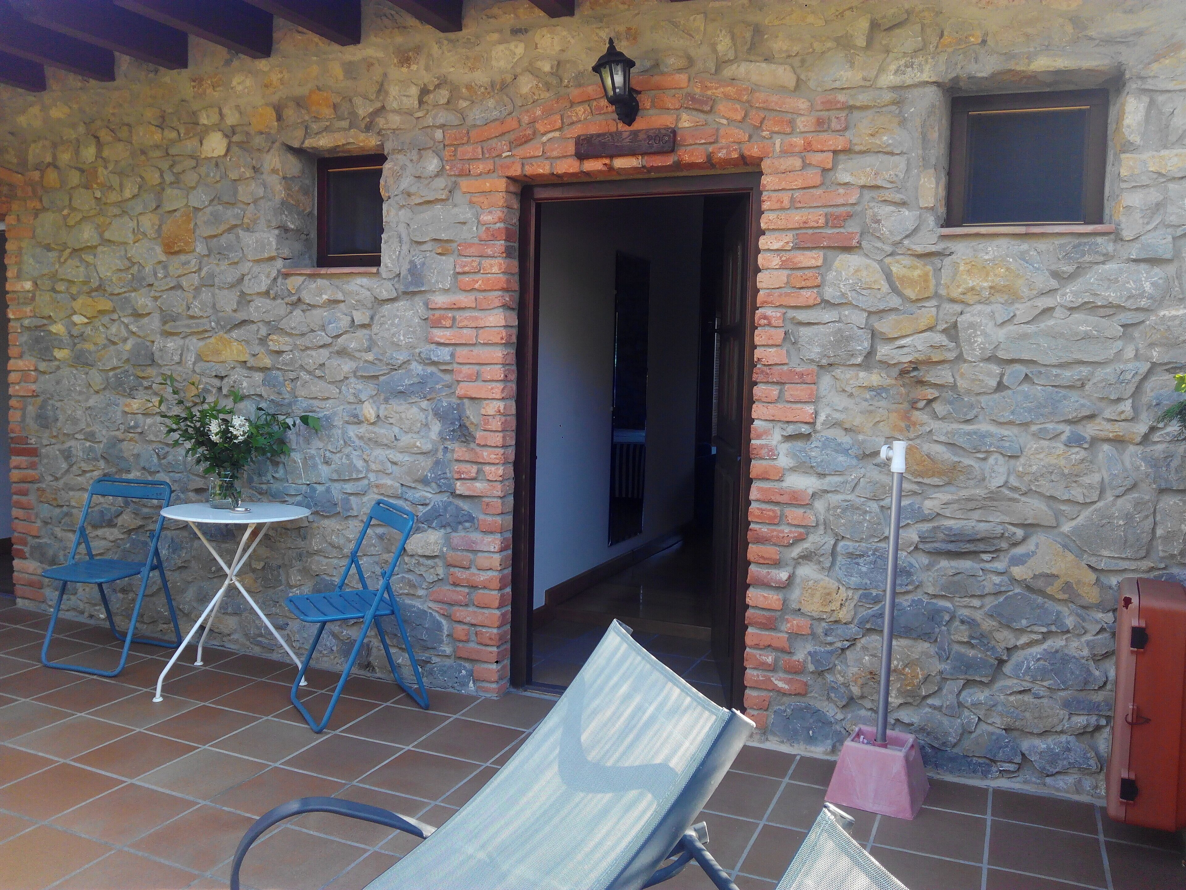 A stone building with a brick archway, two blue chairs, a small table with a vase, and a red suitcase.