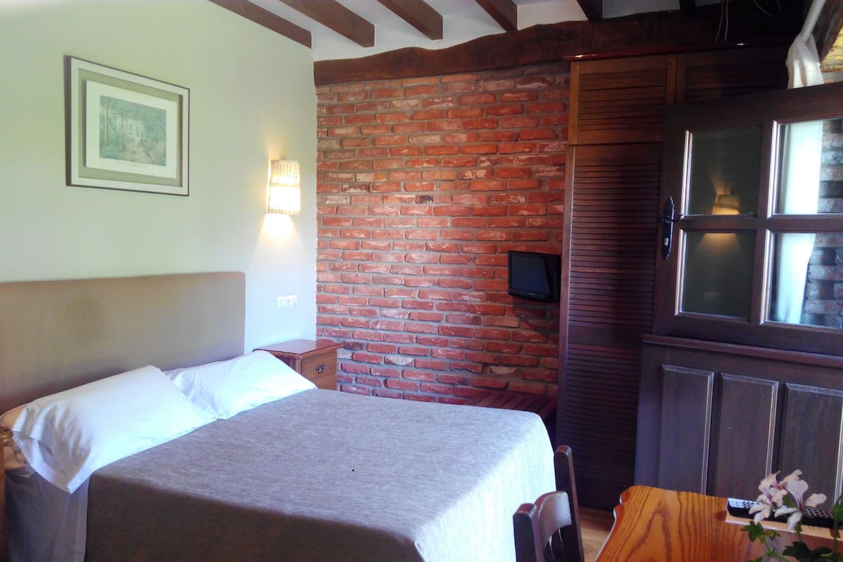 Quarto com cama, mesa de cabeceira de madeira, parede de tijolos, televisão e janela com persianas.