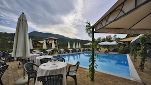 An outdoor pool area with tables and chairs, surrounded by umbrellas and a mountainous backdrop. - Hotel Villa Danilo (Gamberale)