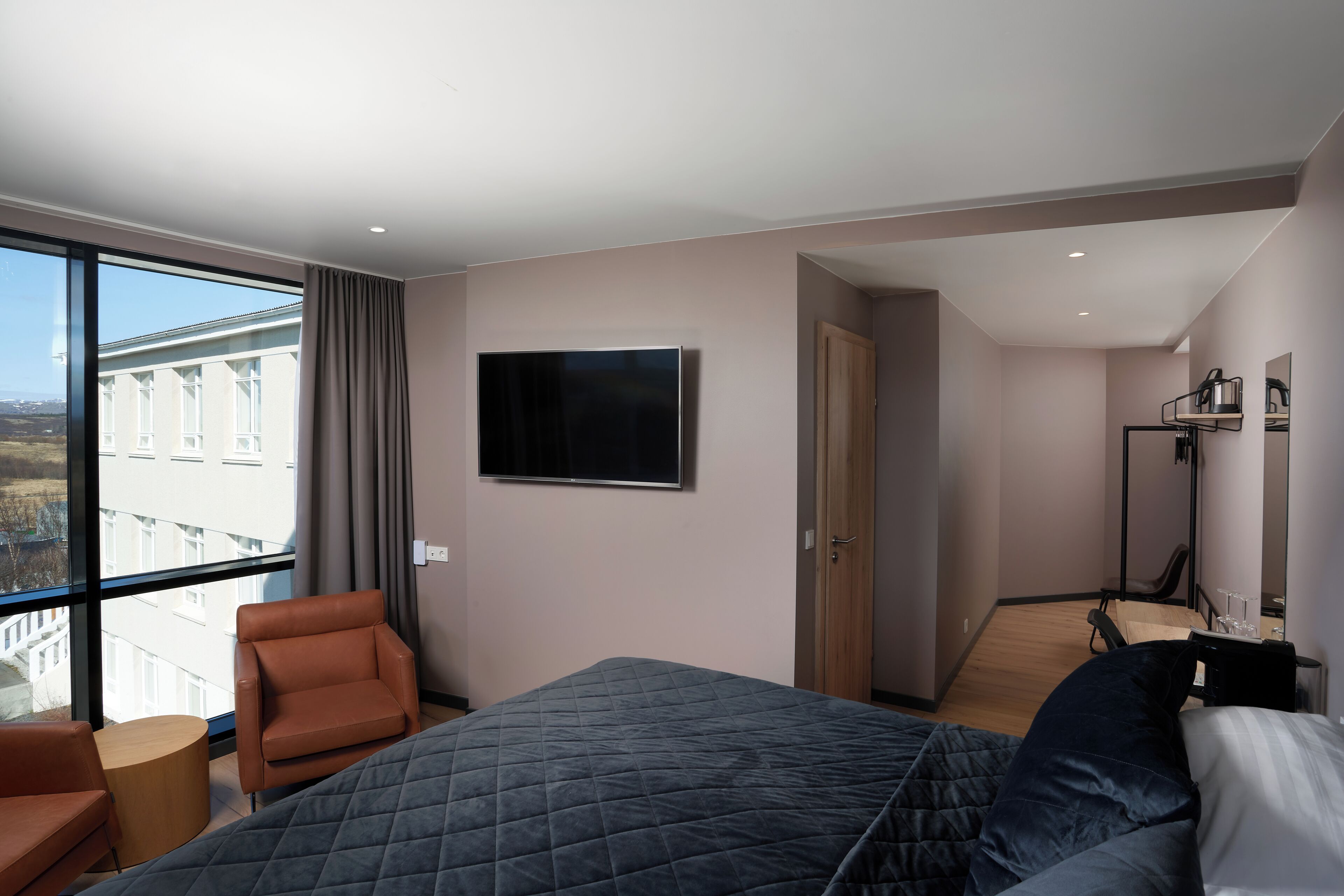 Habitación de hotel moderna con una cama grande, un área de descanso con una silla y una mesita, un televisor de pantalla plana montado en la pared y vistas de la ciudad a través de amplias ventanas.