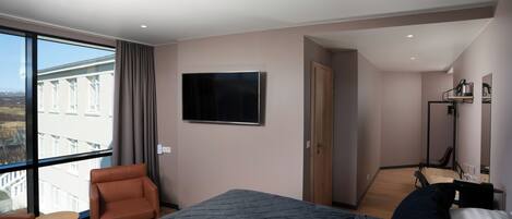 Habitación de hotel moderna con una cama grande, un área de descanso con una silla y una mesita, un televisor de pantalla plana montado en la pared y vistas de la ciudad a través de amplias ventanas.
