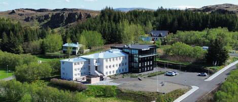 A modern hotel with a flat roof, surrounded by lush greenery and a mountainous backdrop.