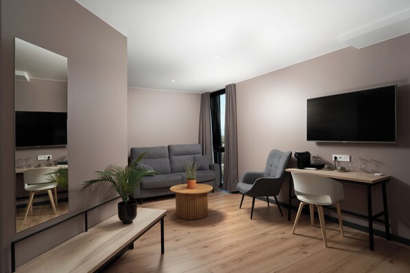 Una sala de estar moderna con un sofá gris, una mesa de centro redonda de madera, una silla blanca, un televisor montado en la pared y un escritorio pequeño con una silla.
