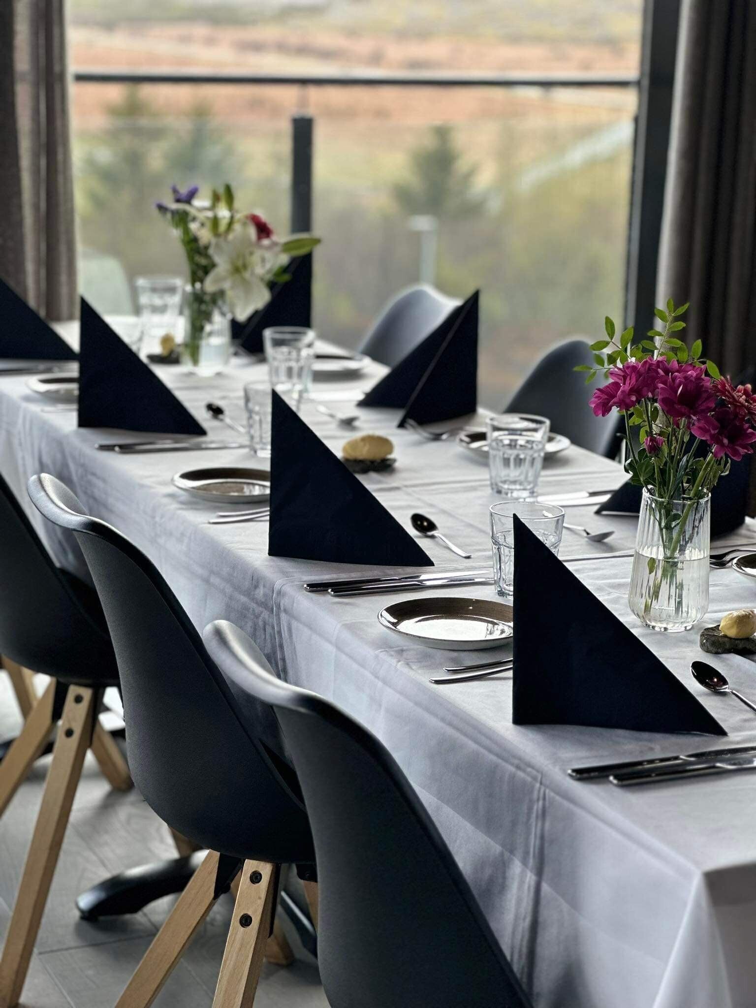 A long dining table set with black napkins, plates, and cutlery, with a vase of purple flowers and a glass of water on the table.