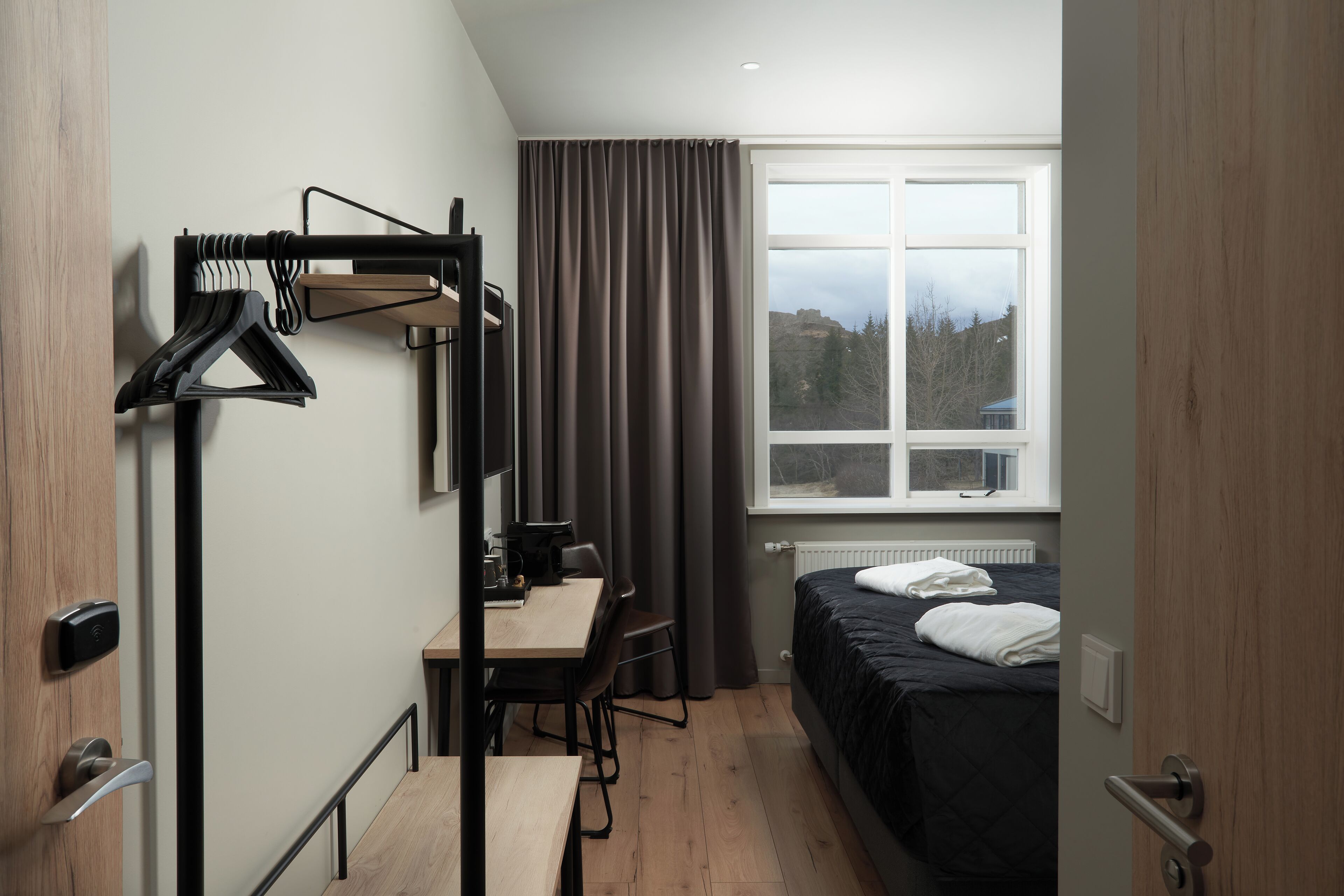 Habitación de hotel pequeña con cama, escritorio y silla. Se ve un perchero con ganchos y una ventana con cortinas.