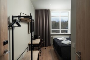 Habitación de hotel pequeña con cama, escritorio y silla. Se ve un perchero con ganchos y una ventana con cortinas.
