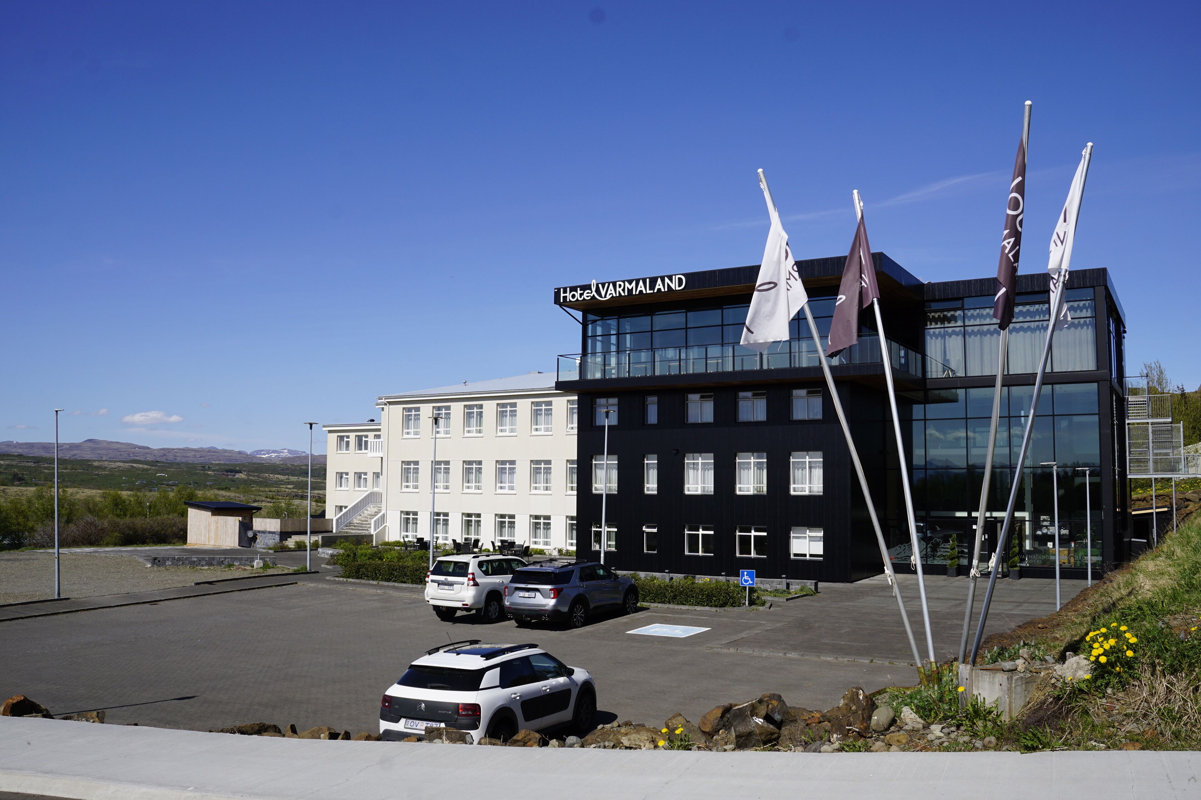 Un bâtiment d’hôtel moderne, avec une enseigne indiquant « Hôtel Varmaland ». On peut voir plusieurs drapeaux à l’avant, un parking avec quelques voitures et un ciel dégagé.