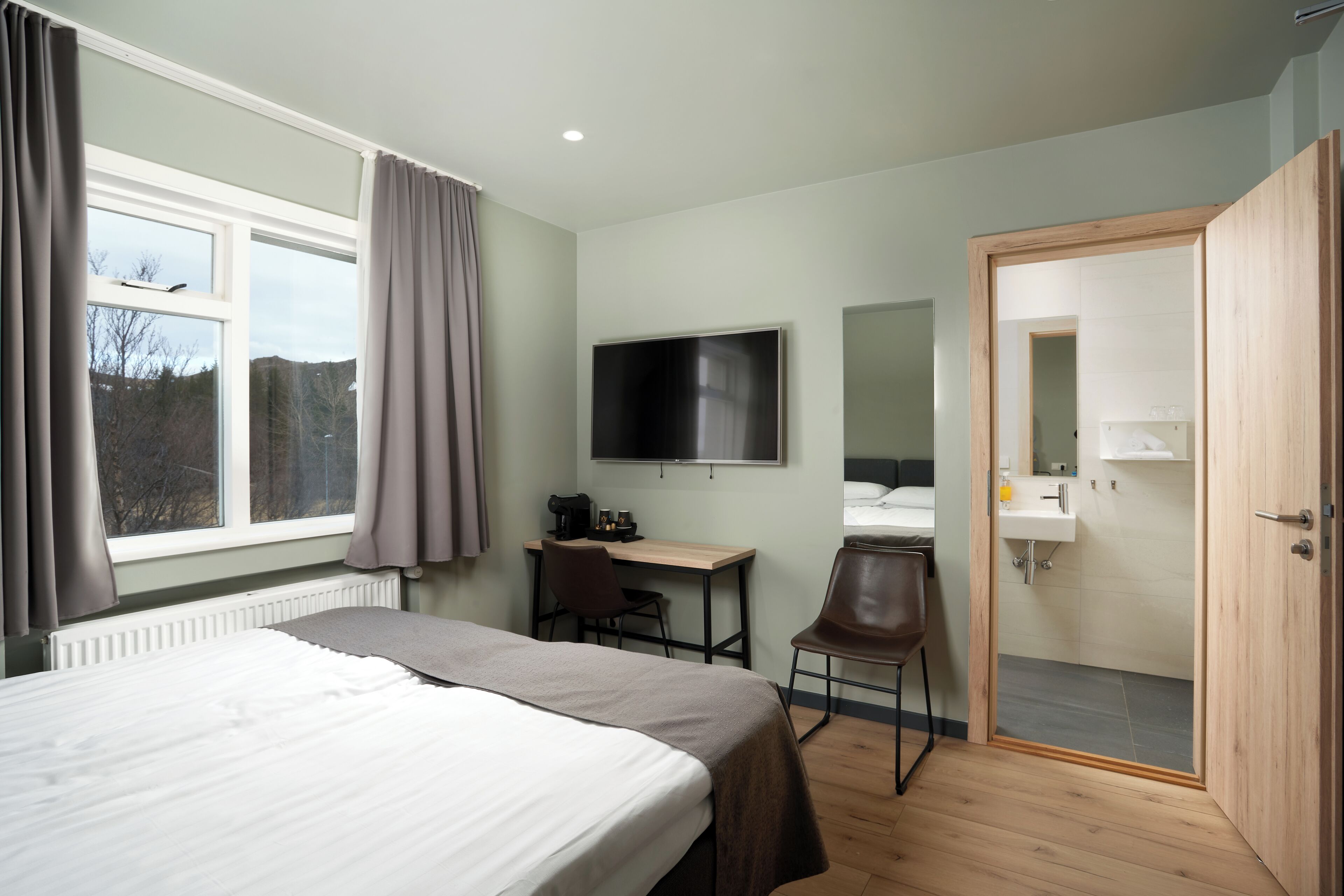 A hotel room with a bed, a desk with a chair, a TV, a bathroom with a sink, and a door.