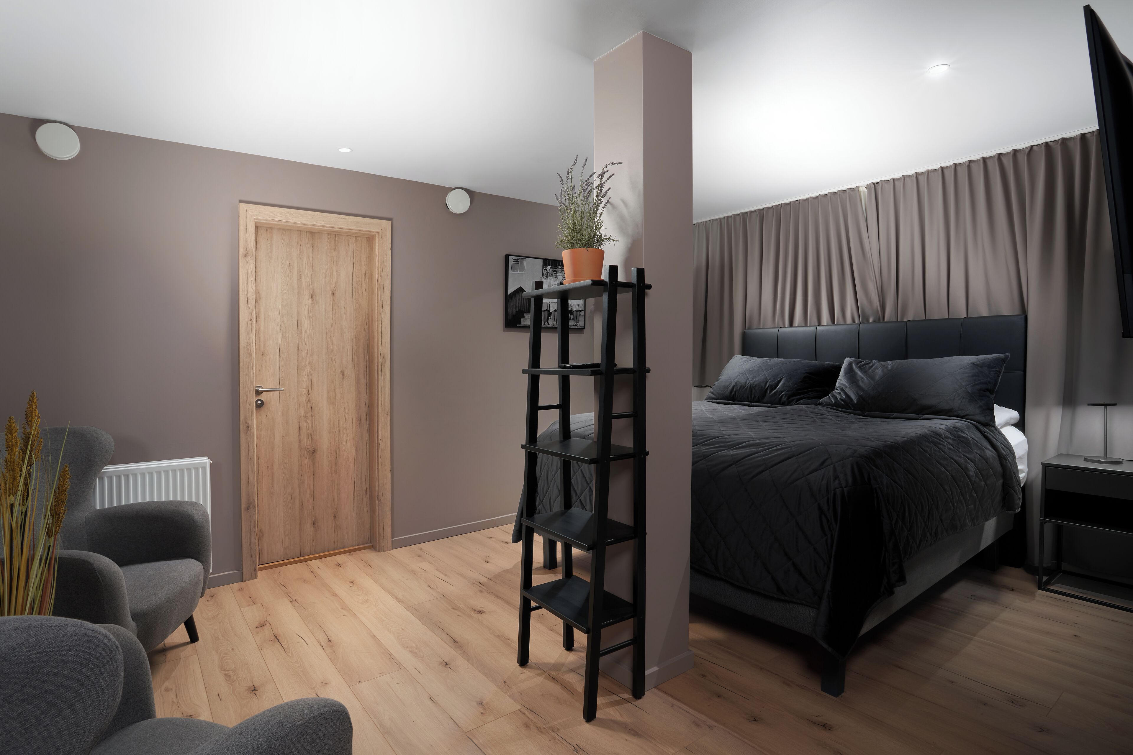 Un dormitorio moderno con una puerta de madera, un estante negro, una cama con ropa de cama oscura y un radiador.