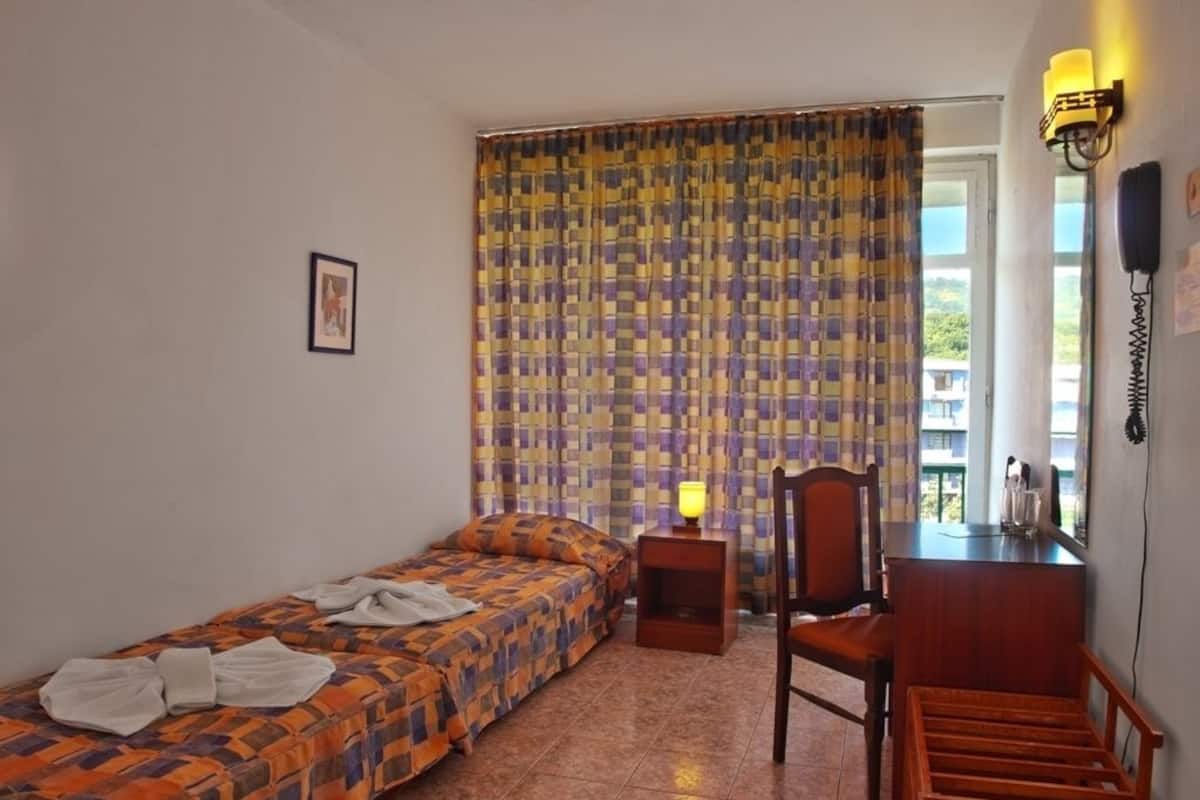 A bedroom with a bed, a chair, a small table, a lamp, and a window with curtains.