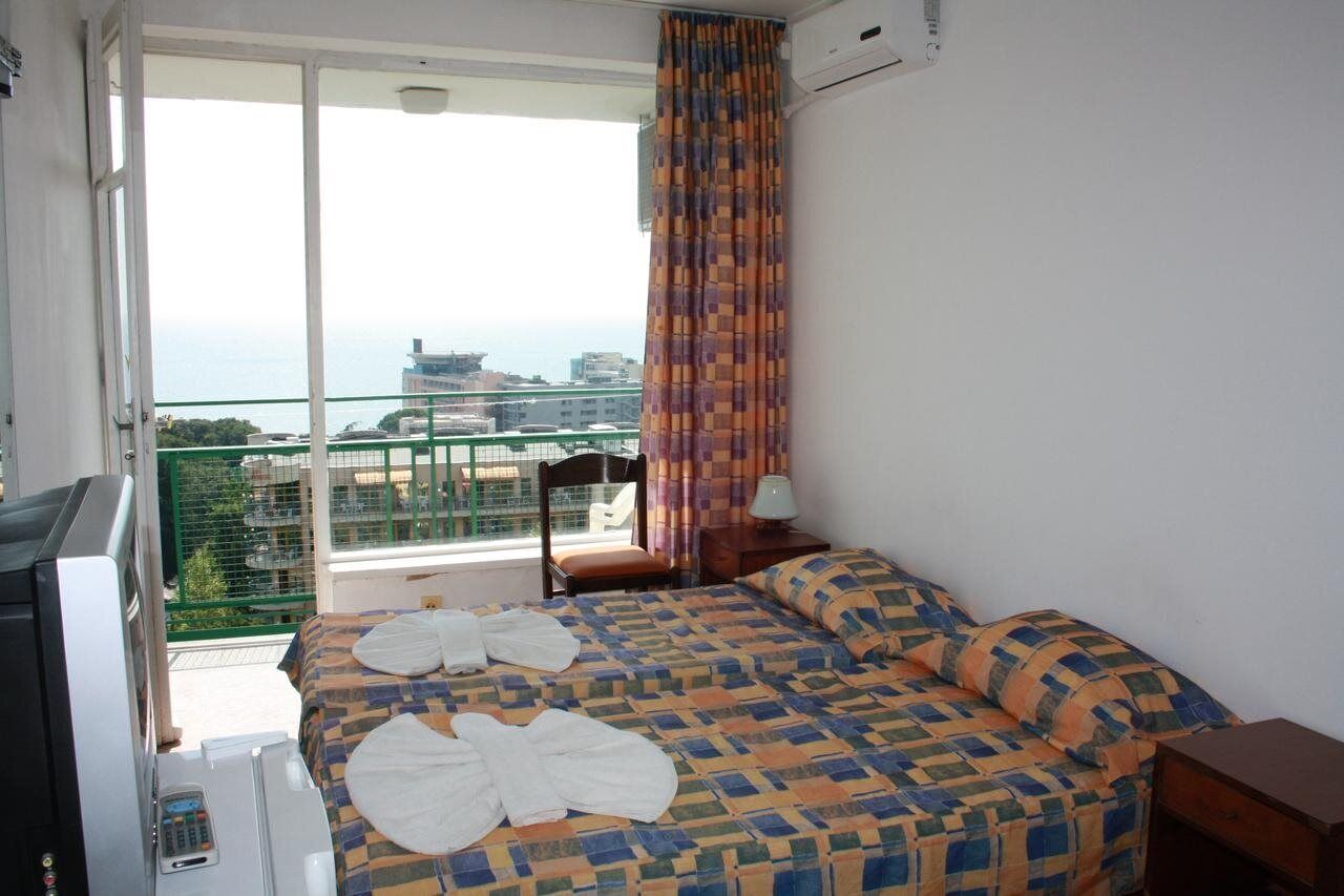 A hotel room with a bed, a chair, a TV, and a balcony with a view of the city and sea.