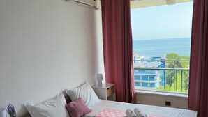 A hotel room with a bed, a nightstand, a window with a view of the sea and buildings, and an air conditioning unit. - Varshava Hotel (Golden Sands)