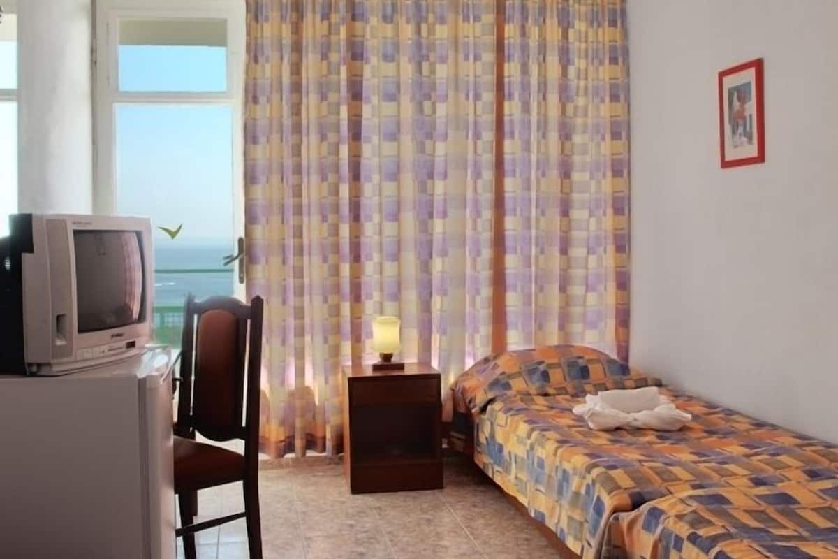A small, tidy room with a bed, a chair, a desk with a television, and a window with curtains.