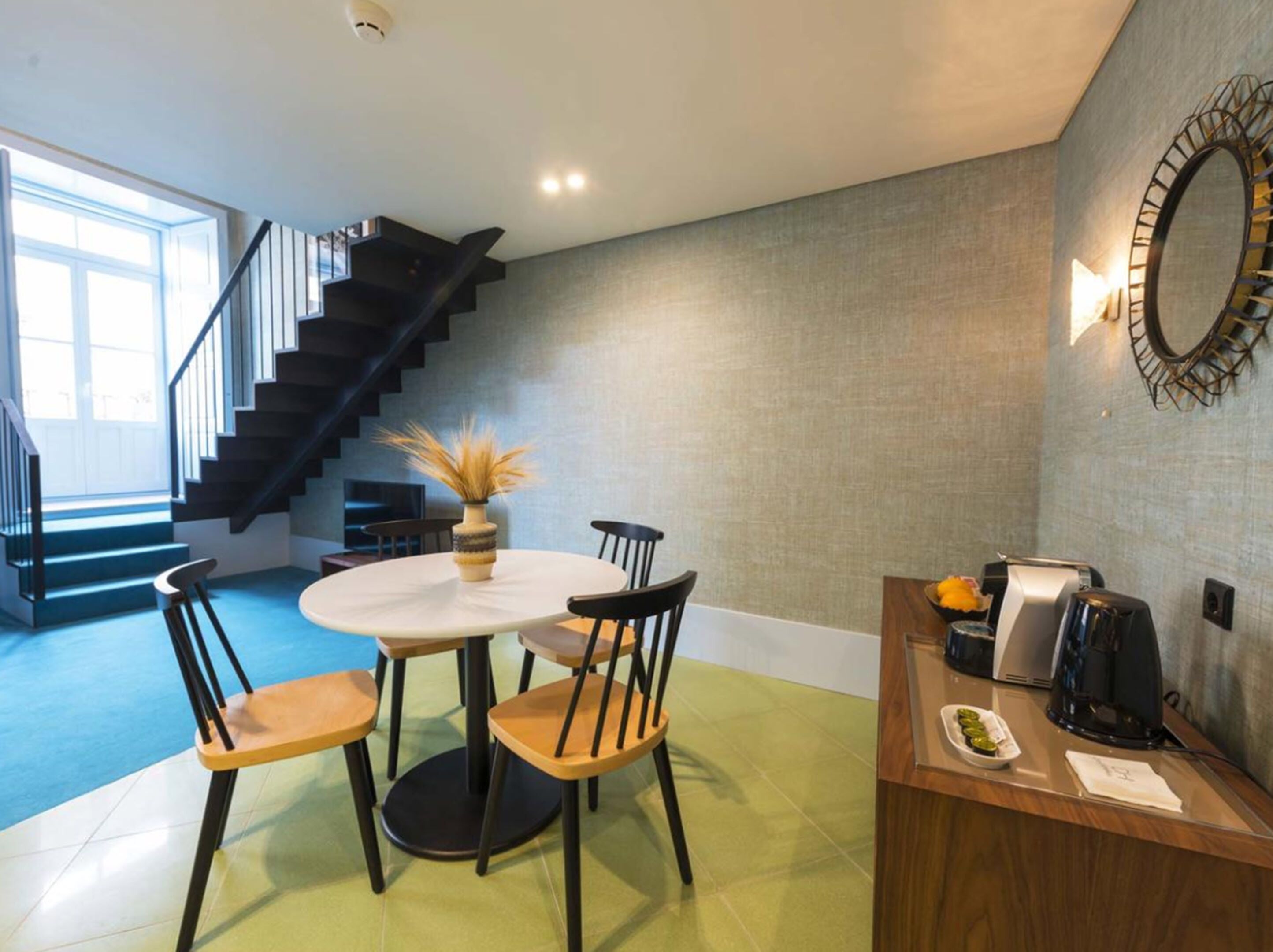 A modern kitchen with a round dining table, four chairs, a coffee machine, and a decorative vase. A staircase is visible in the background.