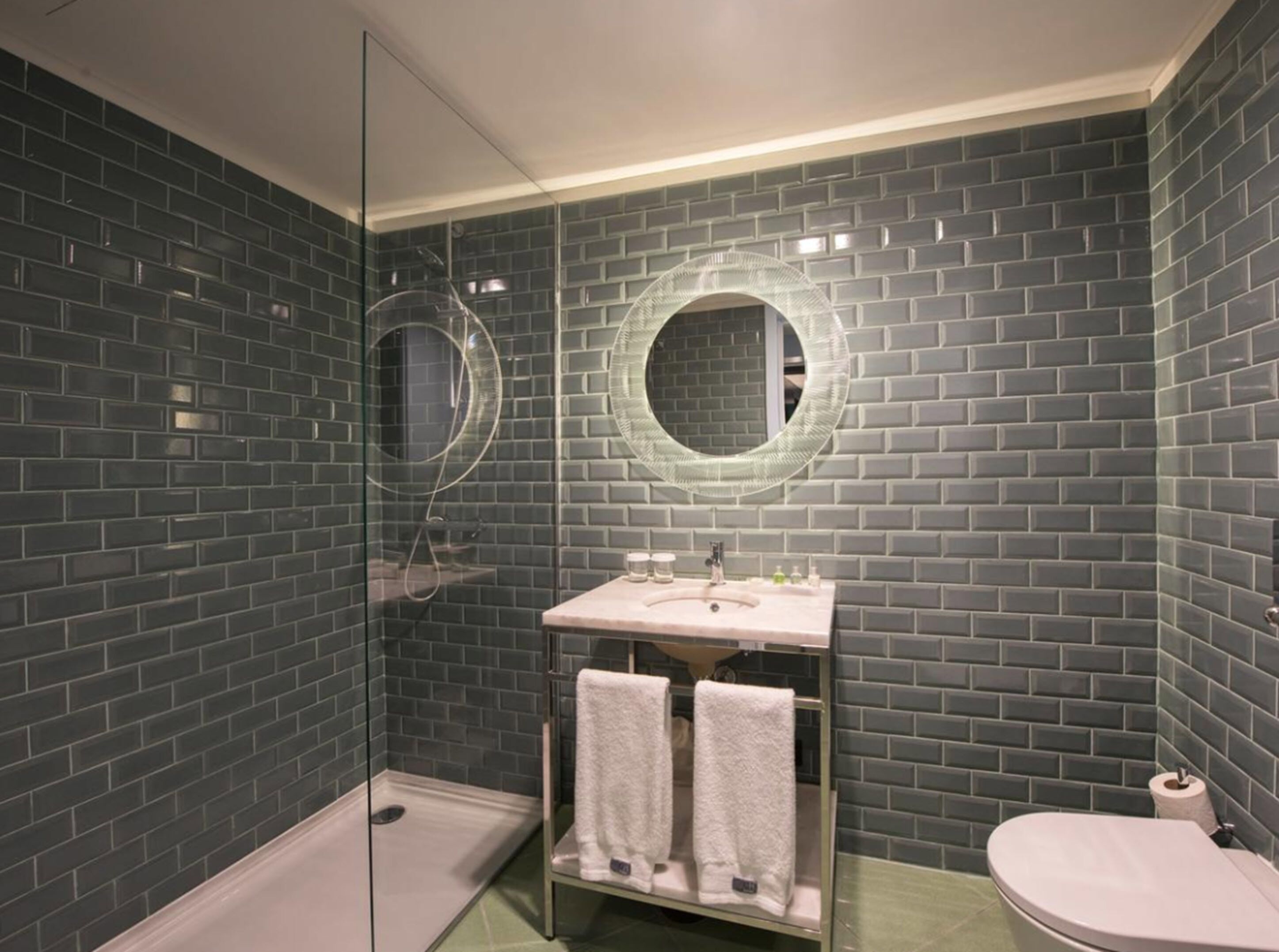 A bathroom with dark gray brick walls, a glass shower enclosure, a sink with a mirror, and a toilet.