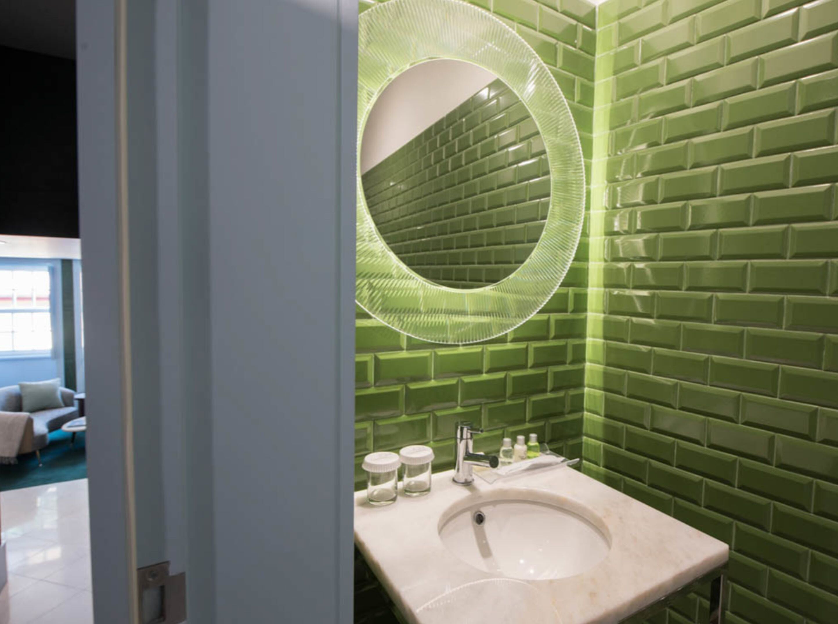 A bathroom with a green tiled wall, a round mirror, and a sink with toiletries.