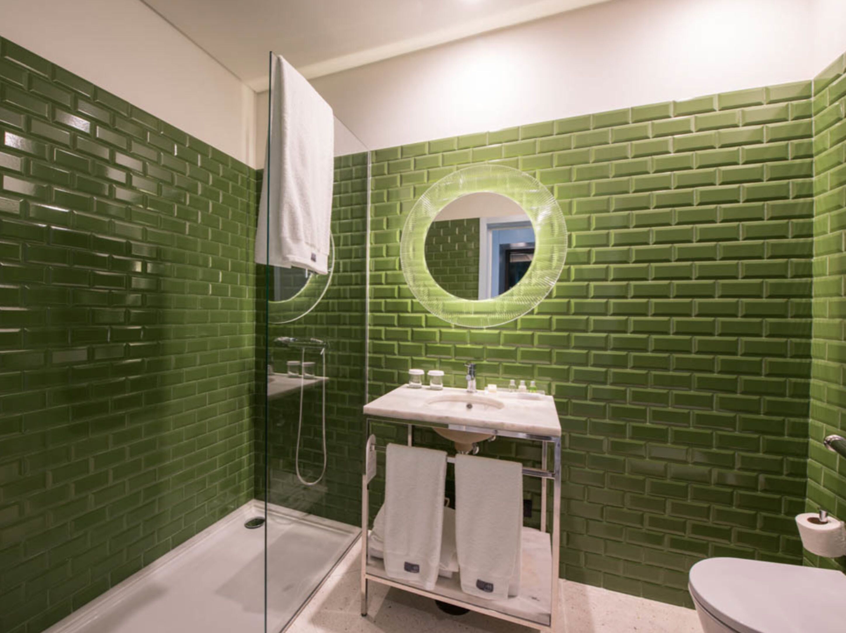 A bathroom with green mosaic tiles, a glass-enclosed shower, a sink with a mirror, and a toilet.