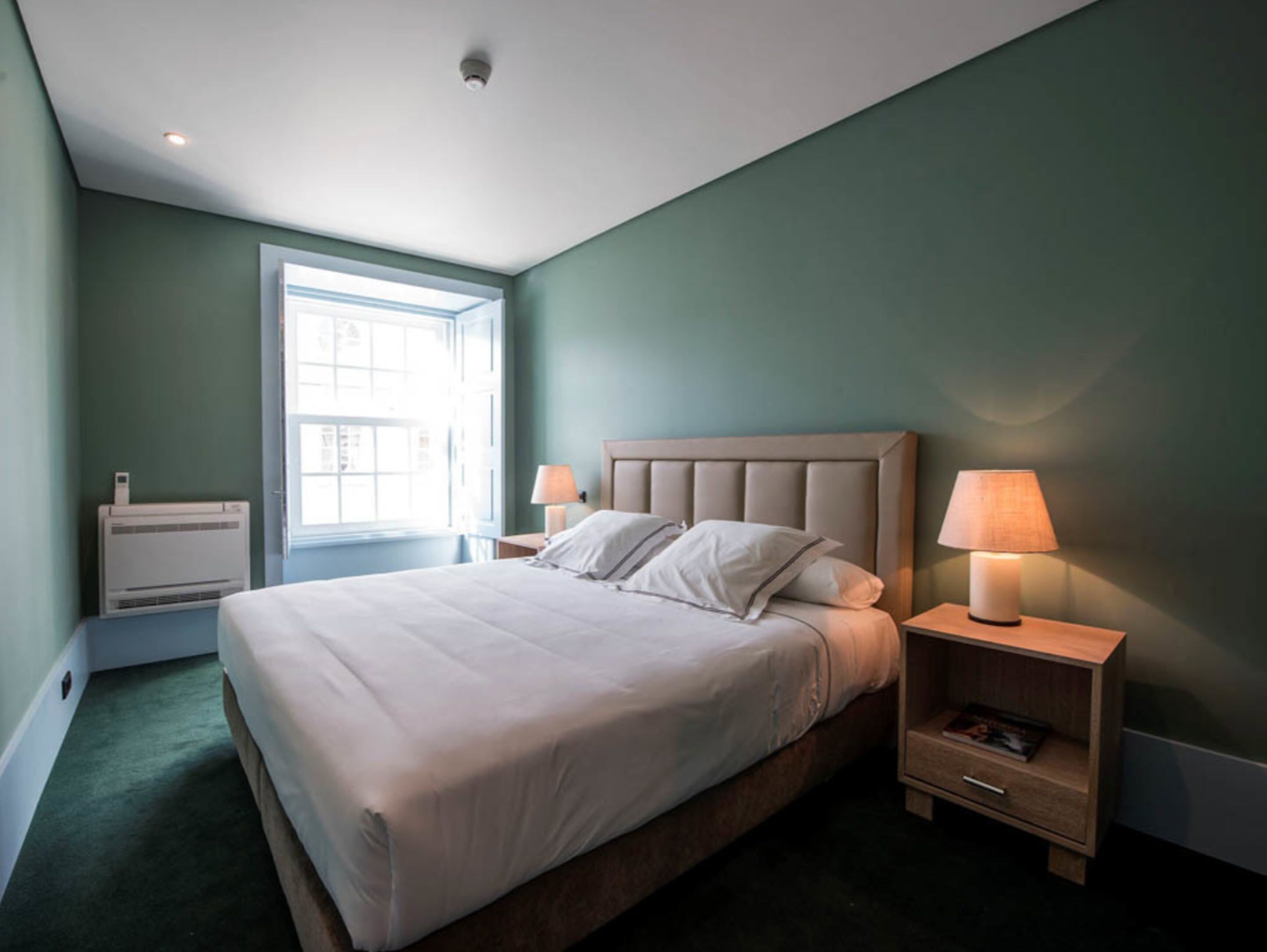 A bedroom with a large bed, two bedside tables with lamps, a window, and a wall-mounted air conditioner.