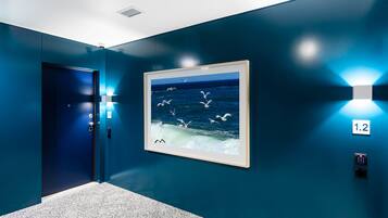 A hallway with teal walls, a framed picture of seagulls flying over water, and a door with a number sign '1.2'.