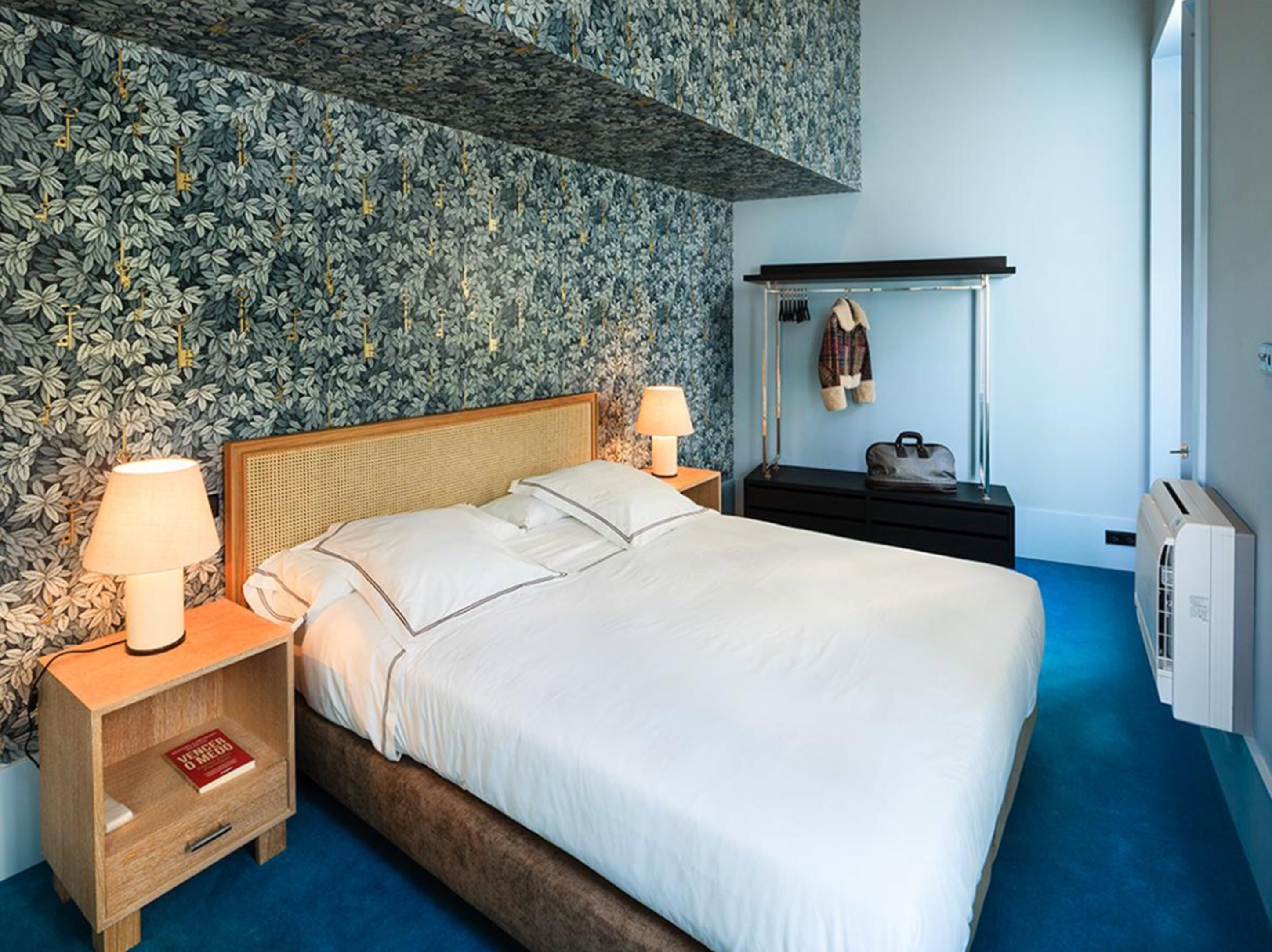 A neatly made bed with white linens, a wooden headboard, and two bedside lamps. There is a small bedside table with a book and a lamp. The room has a blue carpet and a wall with a leafy pattern.