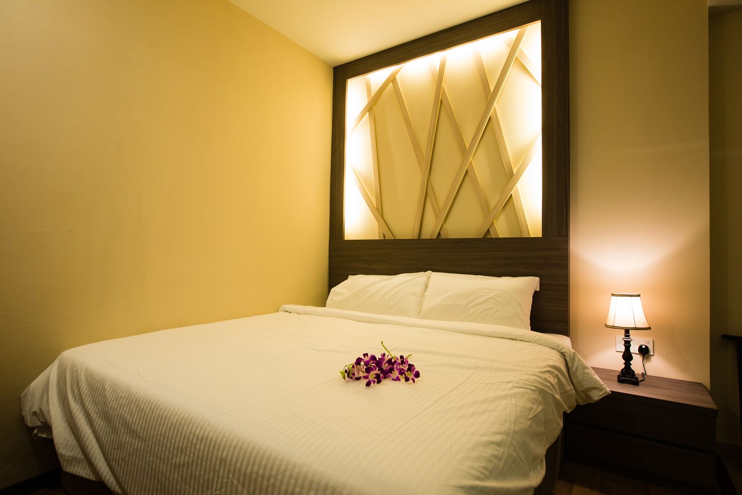A hotel room with a bed, a nightstand with a lamp, and a decorative wall panel.
