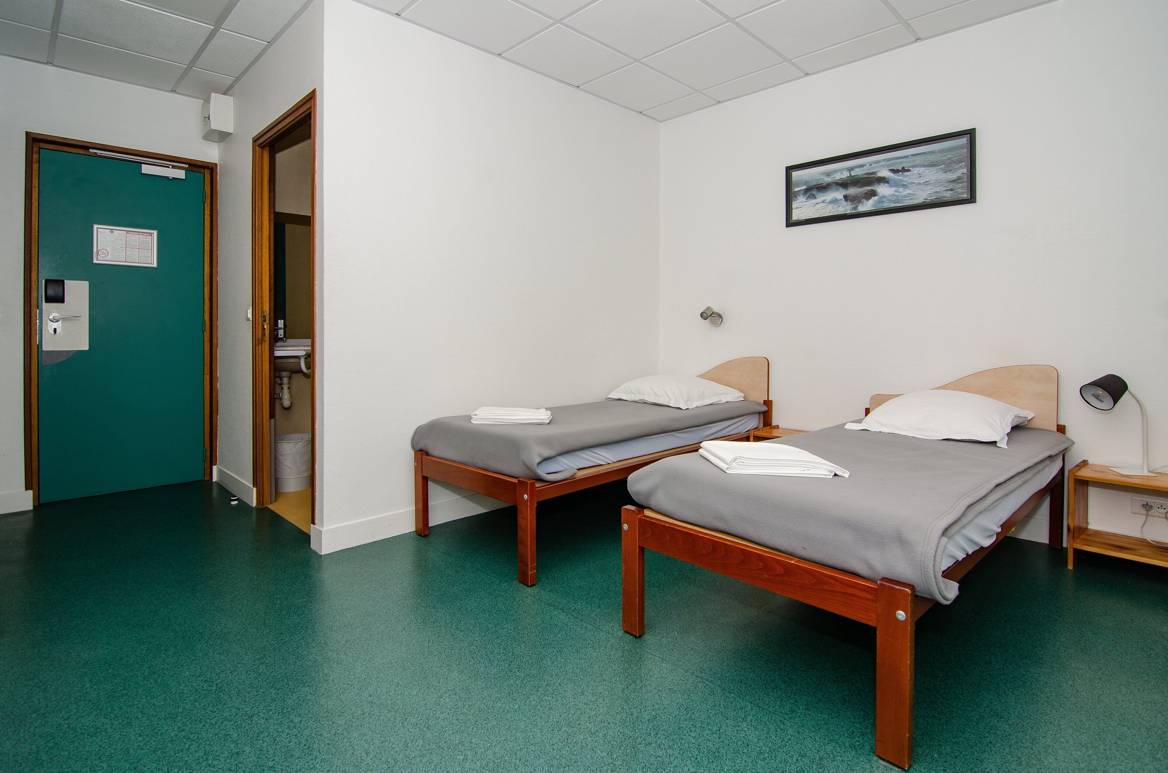 A room with two single beds, a green door, a white wall with a framed picture, and a wooden nightstand.