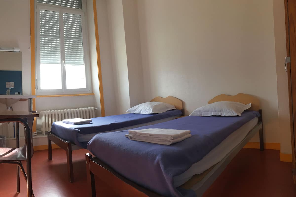 A room with two beds, a desk, a chair, a radiator, and a window with blinds.