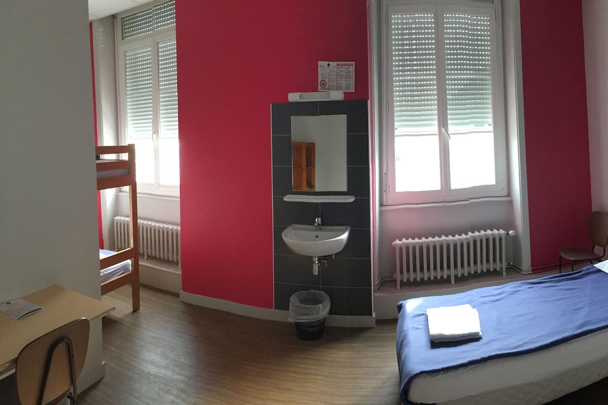 A bedroom with a bed, a desk, a chair, a radiator, and a window with blinds.