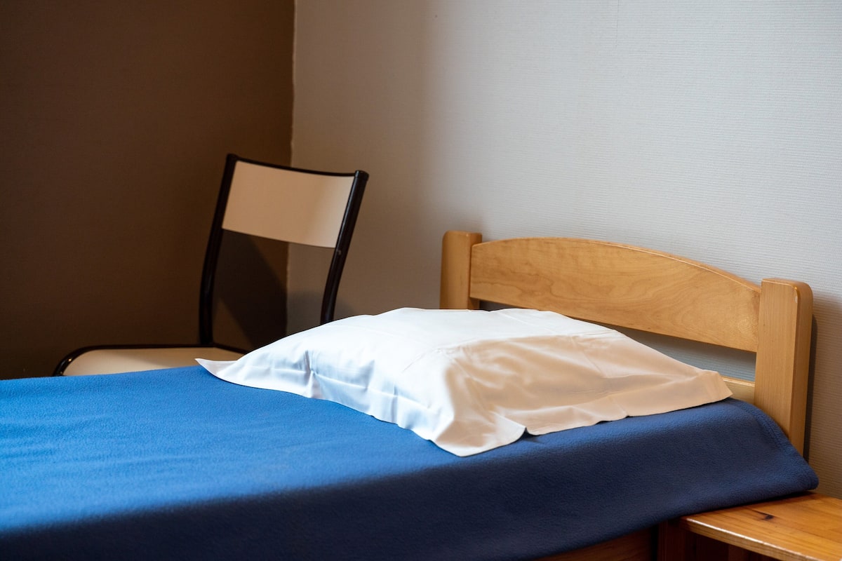 A bed with a blue bedspread and a white pillow, a wooden headboard, and a chair with a black frame.