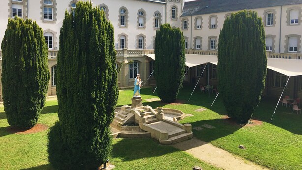 A courtyard with a fountain, trimmed hedges, and a building with multiple windows and a sloped roof.