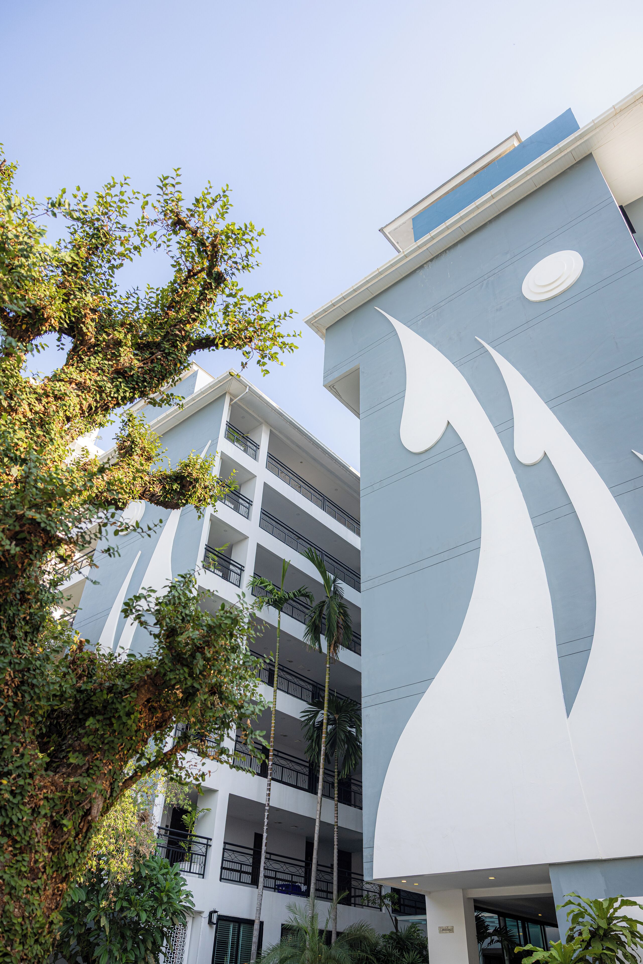 A modern building with a distinctive white and blue facade, multiple balconies, and lush greenery around.