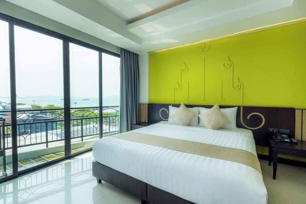 A hotel room with a large bed, a bright green accent wall, a view of the sea, and a balcony with a railing.