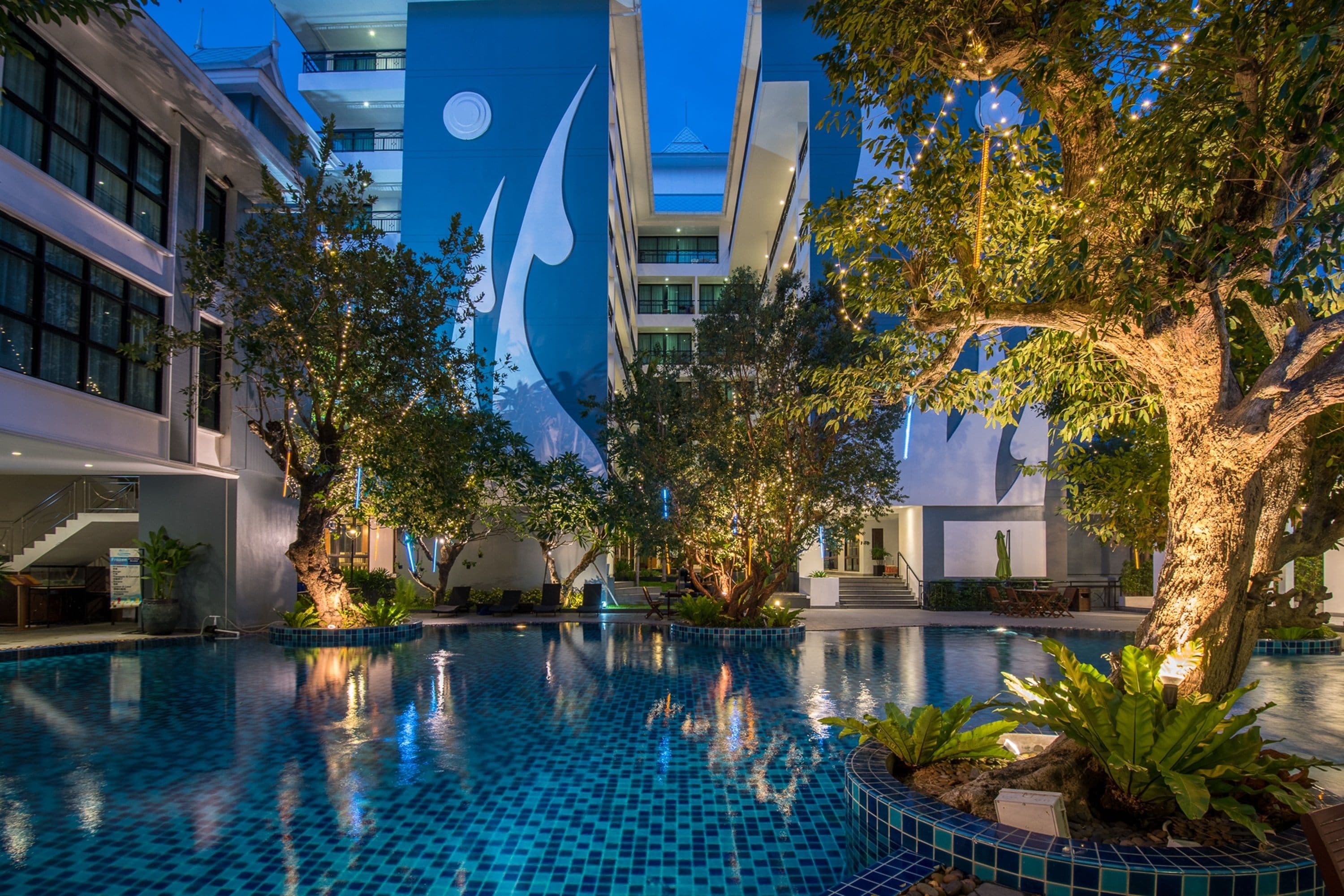 Una piscina interior con suelo de baldosas, rodeada de árboles y edificios modernos con amplias ventanas y un logotipo distintivo azul y blanco.